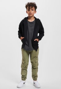 Haut de survêtement zippé noir, t-shirt gris, pantalon cargo vert olive avec poches, et baskets blanches avec accents noirs. Texture de tissu lisse.