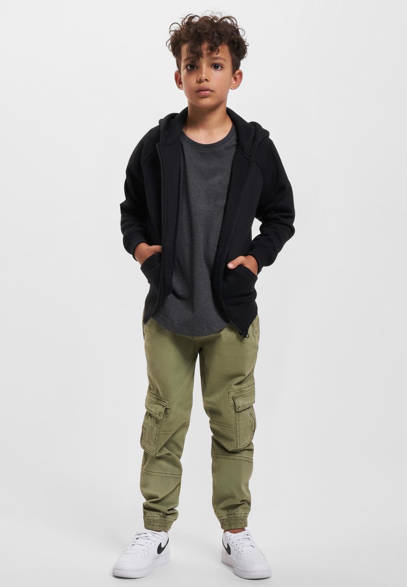 Haut de survêtement zippé noir, t-shirt gris, pantalon cargo vert olive avec poches, et baskets blanches avec accents noirs. Texture de tissu lisse.