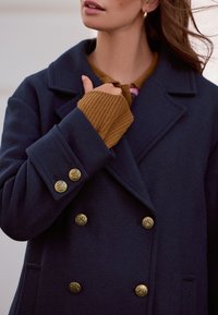 Marineblauwe dubbelde rij jas met gouden knopen en ingezette revers, gedragen over een bruin geribbeld truitje. Materiaal lijkt wolachtig met een gladde textuur.