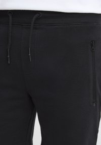 Pantalones deportivos negros con cintura elástica, cordón y bolsillo con cremallera en el lateral; fabricados con un tejido suave y de textura lisa.