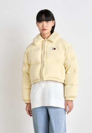 Veste jaune rembourrée, cropped, avec un col doux, une fermeture à boutons et un logo en accent. Portée par-dessus un pull en tricot blanc avec une texture côtelée.
