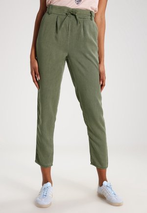 Vrouw die olijfgroene high-waist broek met trekkoord en witte sneakers draagt, staand met handen ontspannen langs haar zij op een effen achtergrond.