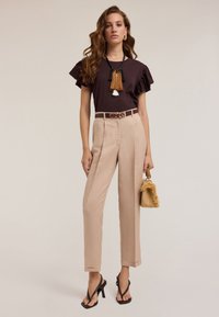 Braunes Top mit rüschierten Ärmeln, beigefarbene, hochtaillierte Hose, schwarze Riemchenheels, dekorativer Gürtel und eine kleine, strukturierte Strohtasche.