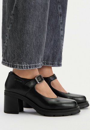 Tacones Mary Jane de cuero negro con hebillas, usados con vaqueros oscuros recortados y pies descalzos.