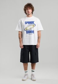 T-shirt blanc à manches courtes avec un texte graphique bleu "SPINNIN' GROOVE." Porté avec un short cargo noir, des baskets blanches et des chaussettes à rayures.