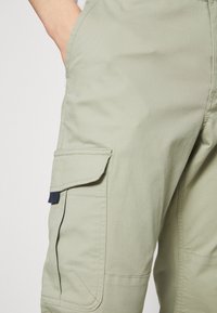 Ljusgröna cargobyxor gjorda av slitstark tyg, med sidofickor och sömnadsdetaljer. Designen inkluderar en marinblå flik på fickan.
