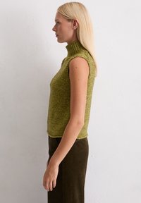 Gebreide mouwloze coltrui in olijfgroen, met een gestructureerde stof en een ribbelkraag. Gecombineerd met een donkergroene corduroy rok.