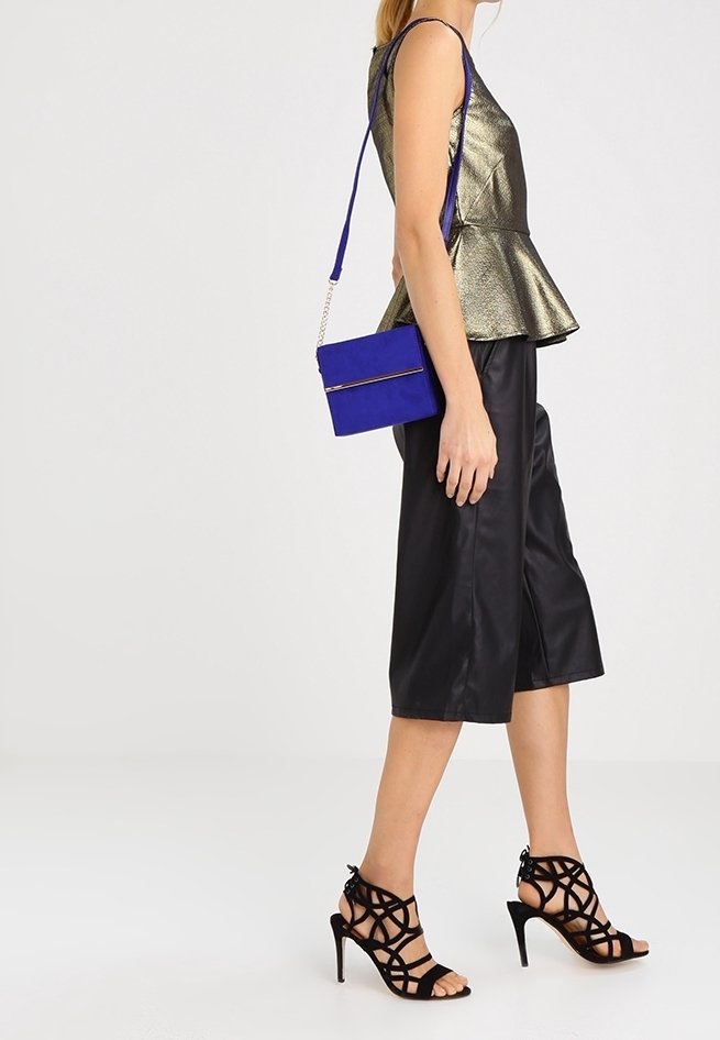 Metallic gold Peplum-Top, schwarze Satin-Culottes, schwarze Riemchen-High-Heels und eine blaue Umhängetasche mit einem goldenen Reißverschlussakzent.