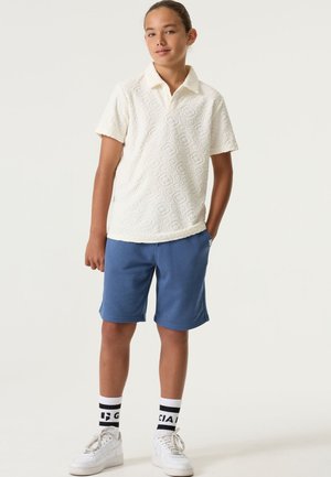 Wit polohemd met textuur en kraag, gecombineerd met blauwe shorts. Het model draagt witte sneakers en sokken met zwarte accenten.