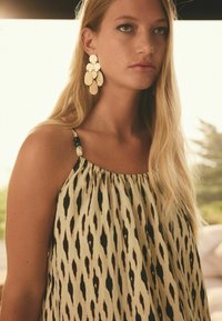 Robe beige avec des motifs ikat noirs, dotée de bretelles spaghetti et de quincaillerie décorative. De grandes boucles d'oreilles dorées accentuent l'ensemble.