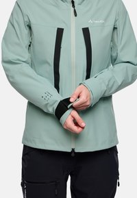 Giacca impermeabile verde chiaro con accenti neri, dotata di tasche sul petto con zip, polsini regolabili e una texture liscia.