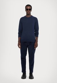 Maglione blu navy a maglia con scollo rotondo, abbinato a pantaloni blu navy coordinati. Il modello indossa occhiali da sole neri e scarpe nere.