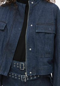 Tumša džinsa jaka ar īsu dizainu, rāvējslēdzēja aizdari, divām priekšējām kabatām un sudraba krāsas metāla detaļām. Kombinēta ar atbilstošu jostu ar acu detaļām.