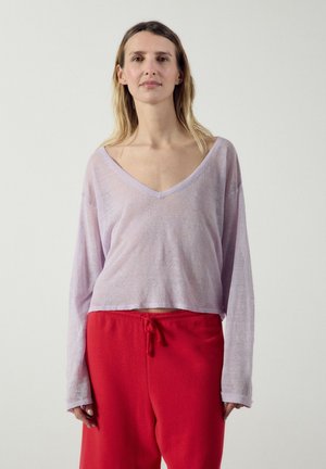 Nainen, jolla on löysä, vaaleanvioletti pitkähihainen paita, jossa syvä V-aukkokohta, ja punaiset kiristysnyörilliset housut, seisoo yksivärisen taustan edessä.