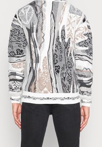 Pull en tricot noir, blanc et beige, avec des motifs tribaux complexes, un tissu texturé et des poignets et un ourlet côtelés.