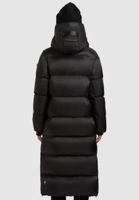 Inimene, kellel on seljas pikk must sulgjope kapuutsi ja must kootud beanie, seistes seljaga lihtsa halli tausta vastu.