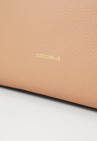 Coccinelle kézitáska puha világosbarna bőrből, texturált felülettel; az oldalon arany-fóliázott logóval rendelkezik.