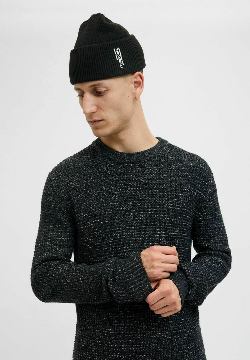 Czarny sweter robiony na drutach z textured wzorem i ściągaczami, w połączeniu z czarną czapką beanie z białą naszywką z logo.