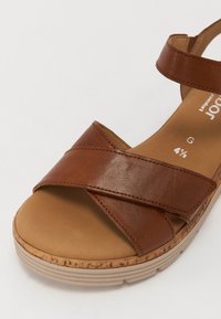 Brown leather sandal with cross strap design, cushioned insole, and beige textured platform sole. Features adjustable ankle strap.