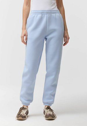 Osoba ubrana w jasnoniebieskie spodnie dresowe z elastycznym paskiem i ściągaczami, w połączeniu z białą górą oraz brązowymi trampkami z białymi sznurówkami.
