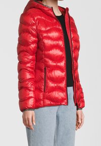 Rød puffjakke med hætte, quiltet mønster, blank tekstur, lynlås fortil og sidelommer. Bæres over en sort top og lyse jeans.
