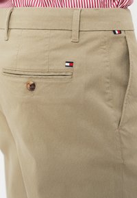 Beige textured trousers with back button pocket and small red, white, and blue Tommy Hilfiger logo, paired with red and white striped shirt.