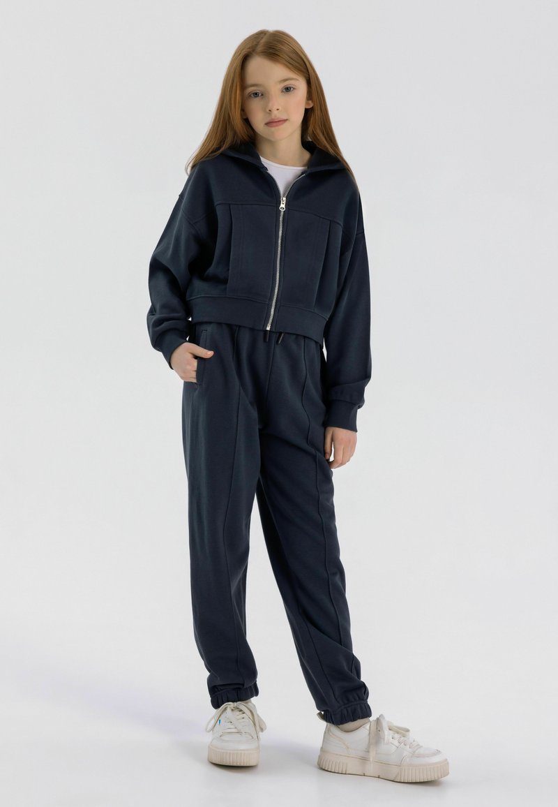 Tmavomodrá skrátená mikina na zips s veľkými vreckami a zodpovedajúcimi jogger nohavicami. Mäkko tkaný materiál s elastickými manžetami. Spárované s bielymi teniskami.