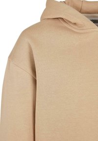 Sudadera con capucha beige que presenta un corte relajado, una textura suave y detalles de costura visibles. La tela parece lisa, adecuada para un uso casual.