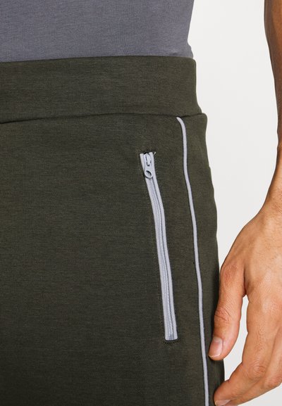 Calças desportivas de um verde escuro com uma textura suave, apresentando um bolso com fecho éclair cinza claro e um acabamento cinza em contraste ao longo do lado.