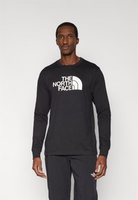 Μαύρο μακρυμάνικο μπλουζάκι με λευκό λογότυπο στο στήθος, στρογγυλή λαιμόκοψη και λεία υφή υφάσματος. Φορεμένο με παντελόνι που ταιριάζει.