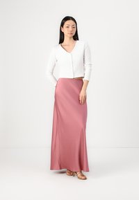 Cardigan court côtelé blanc avec un col en V, associé à une jupe en satin rose longue et fluide et des sandales à talons hauts beiges.