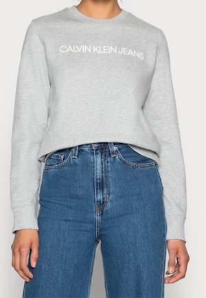 Osoba nosící světle šedou mikinu s dlouhým rukávem s nápisem "CALVIN KLEIN JEANS" a modré džíny s vysokým pasem.