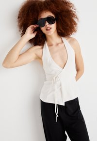 White halter top with a deep V-neck, featuring a drawstring detail. Paired with black relaxed-fit pants. Model wearing oversized black sunglasses.