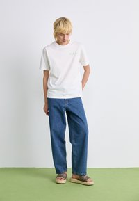 Hvid bomulds-T-shirt med et grønt logo, parret med blå brede jeans og brune mønstrede sandaler med en beige sål, stående på grønne gulve.