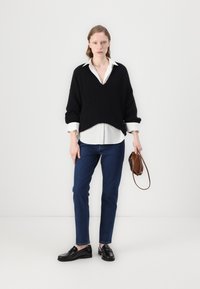Pulover negru supradimensionat cu decolteu în V, cămașă albă dedesubt, blugi bleu și loafers negri. Ține o geantă mică maro.