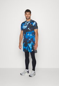 Conjunto deportivo masculino que incluye una camiseta de patrón camuflaje azul, pantalones cortos azules con detalles de tie-dye, mallas negras y zapatillas de correr grises.