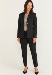 Svart skräddarsydd kostym med en kavaj som har uppslagna slag och två knappar, kombinerad med figurnära byxor och en beige topp. Klassisk och elegant design.