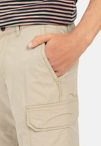 Beige cargoshorts met een zijzak, voorzien van een gladde textuur. Hand rustend in de voortas, wat het functionele ontwerp onthult.
