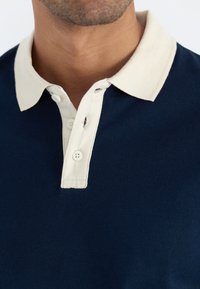 Marineblaues Poloshirt mit einem cremefarbenen Kragen, ausgestattet mit drei Knöpfen und einem kleinen Schlitz am Kragen. Weicher, glatter Stoff. 