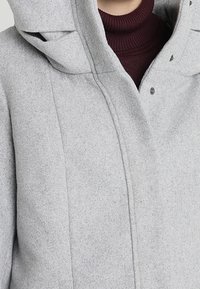 Jasnoszary płaszcz z kapturem zapinany na zatrzaski, noszony na ciemnoczerwonym swetrze z golfem o prążkowanej fakturze.