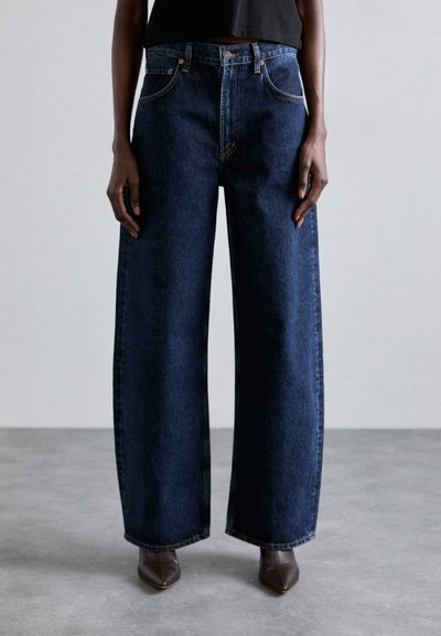 Jeans en denim bleu foncé à jambes larges avec un bouton et une fermeture éclair, cinq poches et des détails de couture subtils. Portés avec des talons pointus marron.