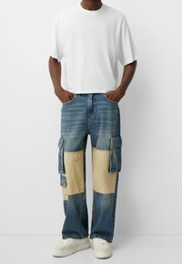 Vit t-shirt med korta ärmar kombinerad med blå denim cargobyxor med beige lappar och flera sidofickor. Tunga sneakers kompletterar outfiten.