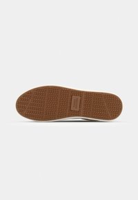 Semelle de chaussure avec une bande de roulement en caoutchouc brun texturé présentant des rainures horizontales et une étiquette rectangulaire centrale avec texte en relief.