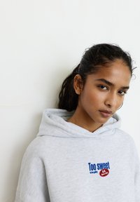 Felpa con cappuccio grigio chiaro con tasca frontale, cappuccio con coulisse e testo ricamato "Troppo dolce per essere colpevole" con un accento a cerchio rosso.