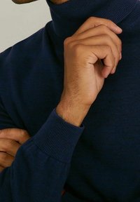 Mörkblå polotröja med ribbstickade muddar och halslinning, tillverkad av texturerat stickat tyg. Händer sammanflätade nära hakan.