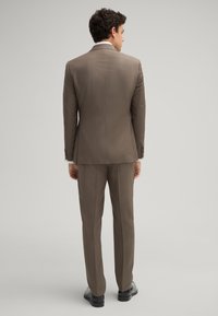 Brauner Anzug mit taillierter Passform, strukturiertem Stoff, einreihigem Sakko und passender Hose. Auffällige Details sind der weiße Hemdkragen und die schwarzen Schuhe.