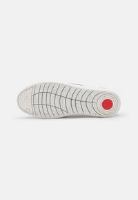 Witte sneakersole met een textuurpatroon, voorzien van een cirkelvormige rode accent en gevlekte details op een flexibel rubbermateriaal.