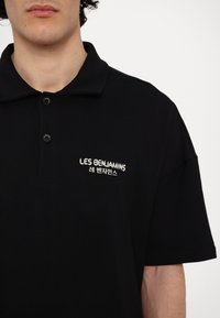 Polo noir à col avec l'inscription « LES BENJAMINS » et des caractères coréens imprimés en blanc sur la poitrine gauche, porté par une personne aux cheveux bouclés.