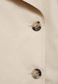 Beige trenchcoattyg med en slät textur, prydd med två stora runda knappar i ett marmorerat mönster och sydda detaljer på sidan.