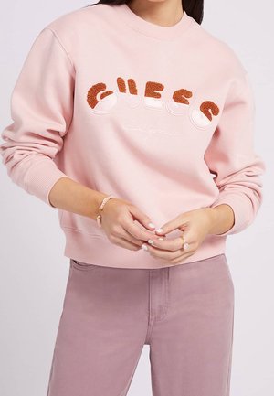Donna che indossa una felpa rosa chiaro con la scritta testurizzata arancione "GUESS" e la scritta "California", abbinata a pantaloni malva e gioielli.